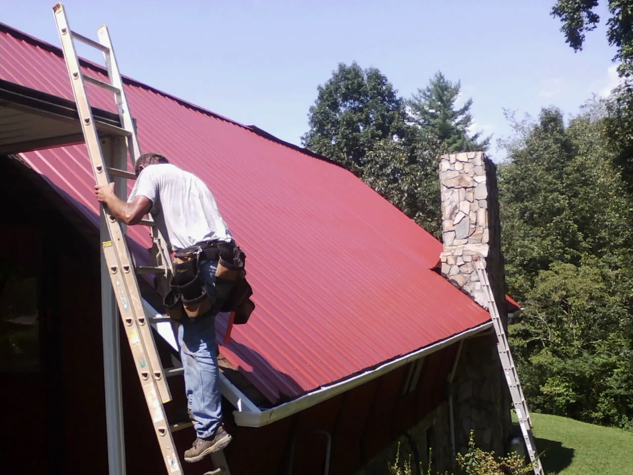 Roofer on a ladder beside a red metal roof and stone chimney — Soffit & Fascia Repair by Roofing Brothers in Adolphus